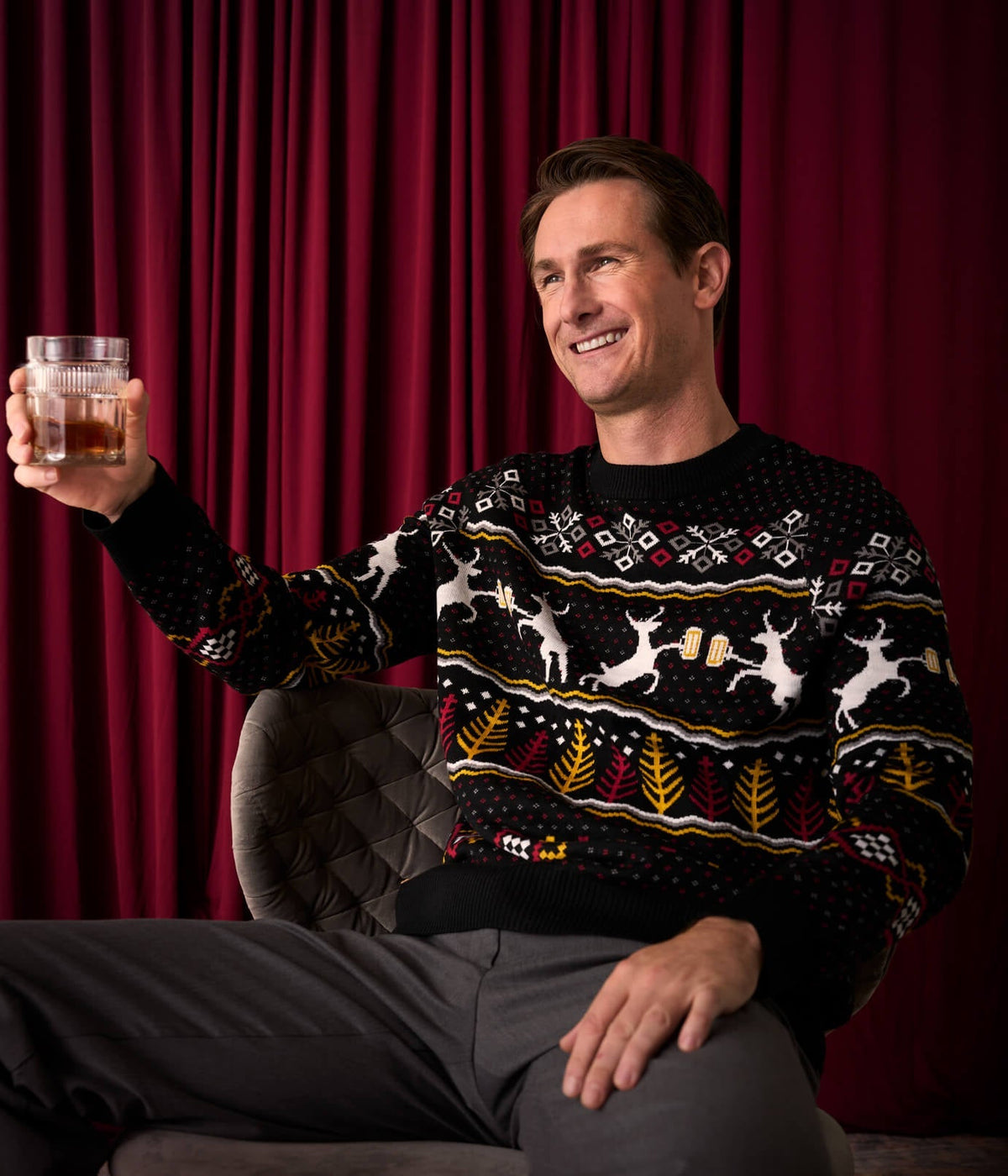 Man wearing a festive sweater holding a glass of whiskey against a red curtain background